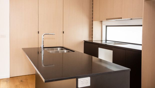 Minimal contemporary kitchen featuring a large dark stone island with integrated double sink and chrome gooseneck tap, pale timber floor-to-ceiling cabinetry, dark lower cabinetry, and a horizontal window above the rear bench. The space is clean-lined and compact with warm natural wood tones and restrained detailing.