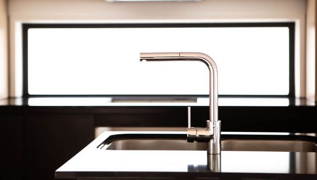 Close-up of a contemporary kitchen sink with a tall polished metal gooseneck mixer tap, dark cabinetry, and a wide horizontal window behind the bench.