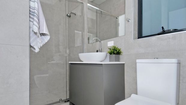 Contemporary compact bathroom with large light-grey tiled walls, a frameless glass shower enclosure, white vessel basin on grey vanity cabinetry, chrome tapware, wall mirror, and a white toilet. A small plant and folded towel add modest decorative detail.