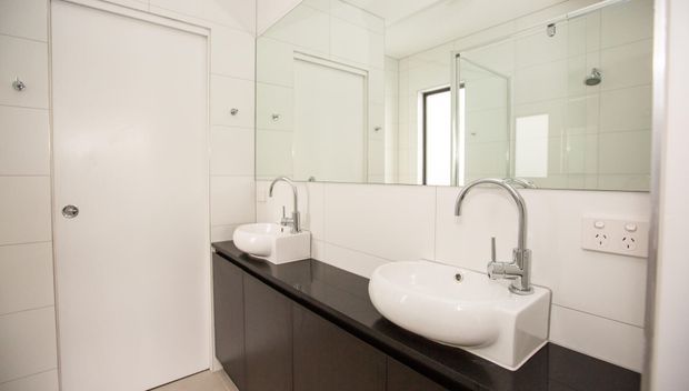 Bright contemporary bathroom with twin white vessel basins on a dark vanity, curved chrome tapware, large wall mirror, white tiled walls, and a glass-enclosed shower visible in the reflection.