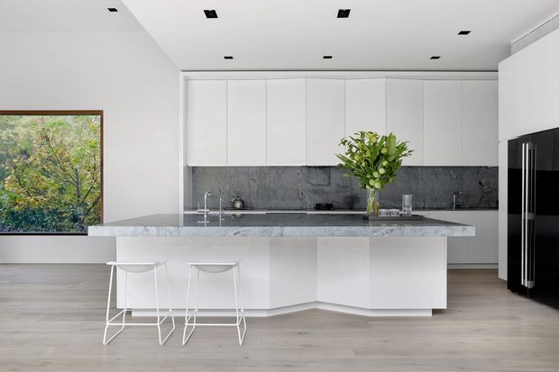 Bright contemporary kitchen with a large white island and waterfall-style stone benchtop, integrated white cabinetry, dark textured splashback, black appliances, pale timber flooring, and two minimalist white bar stools. A framed garden view is visible through the window.