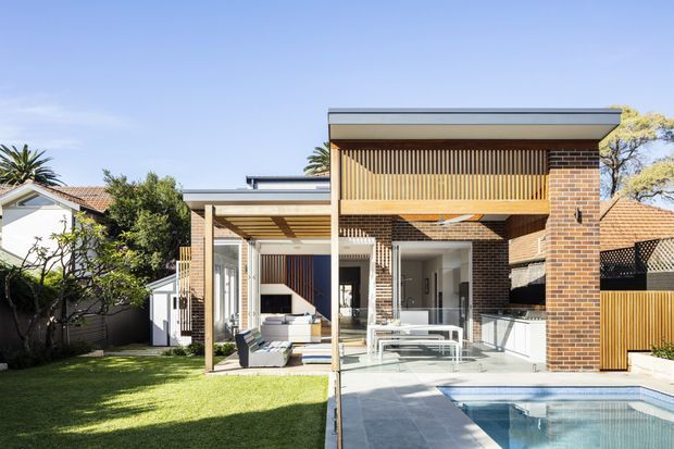 Bright exterior view of a contemporary two-storey brick house with a flat roof, timber batten screening, extensive glazing, a covered outdoor entertaining area, paved terrace and swimming pool. The design opens the living spaces toward a landscaped backyard and pool, with neighbouring residential buildings visible around it.