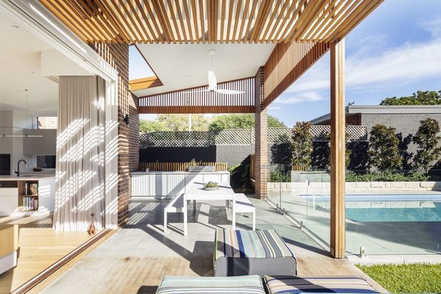 Bright contemporary indoor-outdoor living area opening onto a pool terrace. A timber slatted pergola and matching overhead screens create strong linear shadows over a concrete-paved dining zone with a white table and chairs. The open kitchen and living interior are visible at left, while a fenced swimming pool and landscaped courtyard sit beyond the glazing. Brick piers, timber posts, and extensive outdoor connection define the composition.