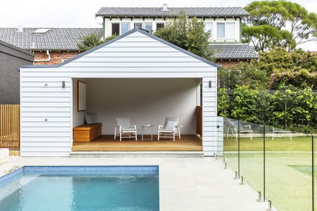 A contemporary backyard pool area with a freestanding, gable-roofed pavilion or pool house clad in pale horizontal weatherboards. The open-fronted structure contains a timber-floored outdoor seating area with two lounge chairs, a small table and built-in timber storage. A glass pool safety fence encloses the blue swimming pool and adjacent lawn, while a larger brick-and-rendered tiled-roof house is visible behind.