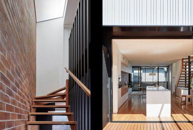 A split architectural view of a contemporary multi-level home. The left side shows an open timber staircase beside an exposed red-brick wall and black vertical metal screening, with a skylit stairwell. The right side looks through a dark-framed opening into a bright open-plan kitchen and dining area with timber flooring, a white island, extensive glazing, and a timber stair or screen element.