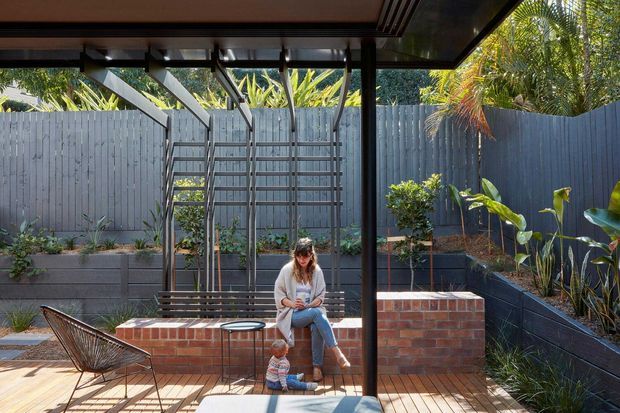 A covered outdoor courtyard or patio viewed from beneath a modern roof structure. The space features a timber-look deck, dark metal posts and beams, a brick built-in bench or planter wall, raised garden beds, grey vertical fencing, and a timber trellis. Tropical planting surrounds a seated adult and child, with strong sunlight and shadows creating a warm, relaxed atmosphere.