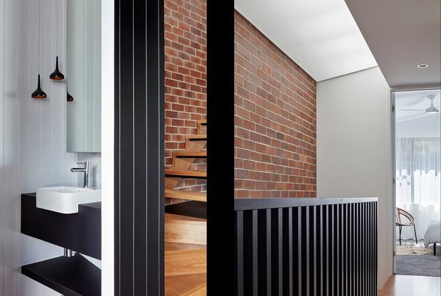 Interior view of a contemporary multi-level home featuring a compact bathroom vanity at left, a timber staircase with black metal detailing, and an exposed red brick feature wall. Vertical black battens form a stair balustrade and screen, with a glimpse of a bright living or dining area beyond.