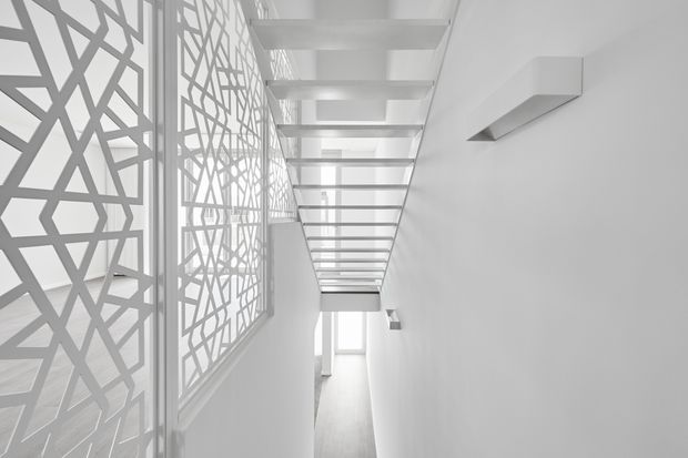 A bright, narrow contemporary passage or stair hall in a predominantly white interior. The left side features large patterned geometric privacy screens over glazing, while the ceiling has a long sequence of exposed horizontal beams or slats creating strong linear perspective. Minimal wall-mounted rectangular light fittings and a bright window at the far end emphasize the clean, monochromatic architectural design.
