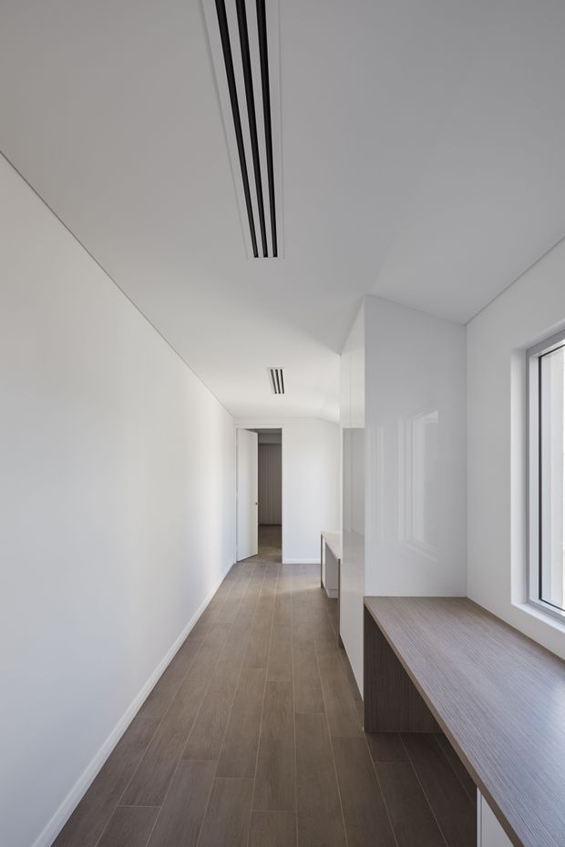 A narrow, bright minimalist hallway with white walls and ceiling, recessed linear air vents, warm grey-brown plank flooring, and a long built-in timber-toned window bench or console beneath a large window. A doorway opens to another space at the far end. The design is clean and highly pared back, with strong natural light and integrated joinery.