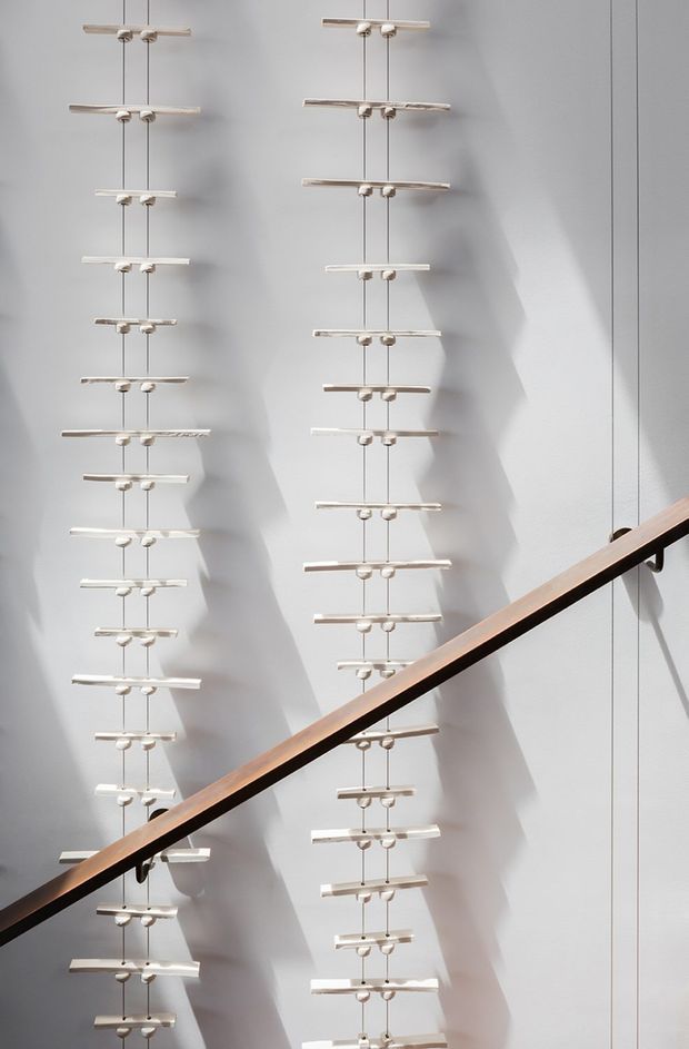 Vertical interior view of a tall stairway or landing with two repeated hanging sculptural light installations suspended against a pale wall. A dark timber handrail cuts diagonally across the foreground, while strong daylight creates elongated shadows from the fixtures. The composition is minimalist and architectural, emphasizing the double-height volume and linear pendant arrangement.