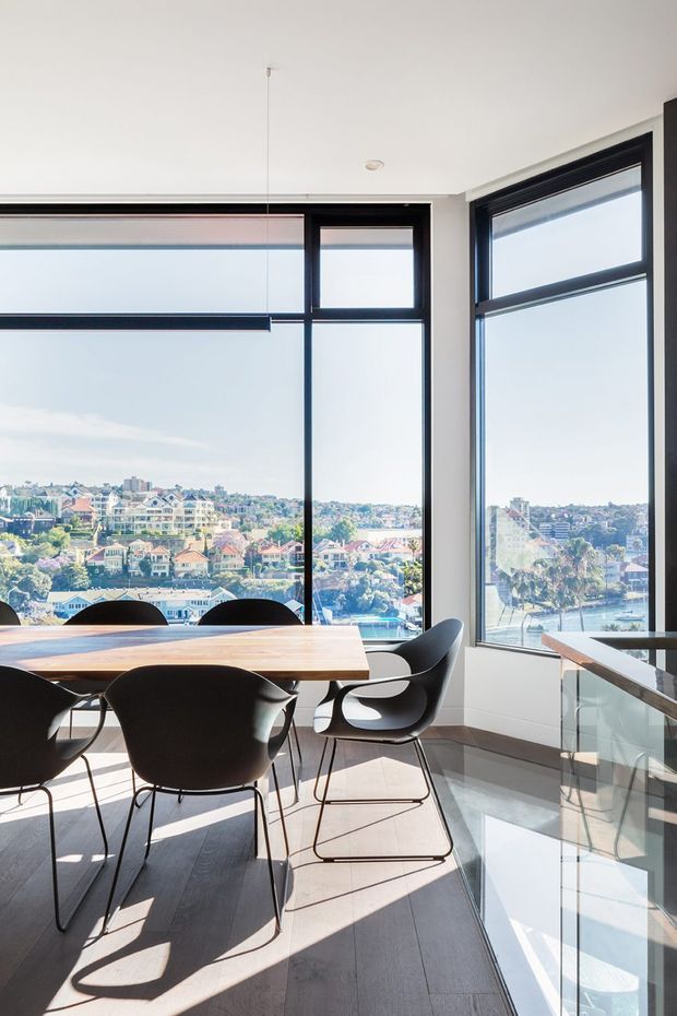 Bright contemporary dining area with a rectangular timber dining table and sculptural black chairs positioned beside expansive black-framed corner glazing. The room has pale walls, dark timber flooring, a glass balustrade or partition at right, and broad views over a dense residential waterfront or harbour-side neighbourhood. Strong daylight and reflective surfaces create an airy, open atmosphere.