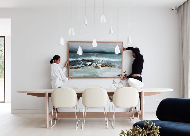 Bright contemporary dining area with a pale timber dining table and three upholstered white chairs, beneath a cluster of sculptural white pendant lights. A large ocean-themed artwork forms a feature above the table, while sheer curtains, light walls and pale flooring create an airy setting. A portion of a dark sofa is visible in the foreground, and people are adjusting the artwork and table styling.