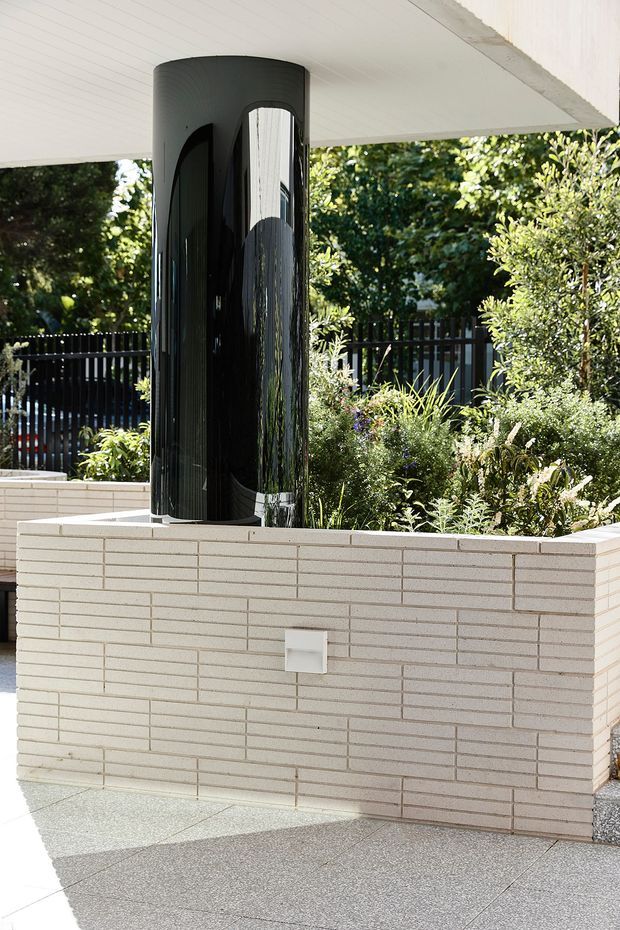 Close-up of a contemporary outdoor area featuring a glossy black cylindrical structural column supporting a white soffit, set behind a pale brick planter wall filled with dense landscaping. A small square wall light is integrated into the planter, and the ground is finished in light grey textured paving.