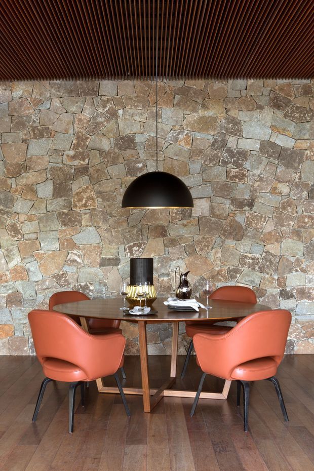 A compact dining area with a round dark timber dining table and four sculptural rust-orange upholstered chairs. The room features a rugged irregular natural-stone feature wall, warm timber flooring, a dark timber slatted ceiling, and a large black dome pendant light. Tableware and decorative objects are arranged on the table, creating a warm, refined, earthy atmosphere.