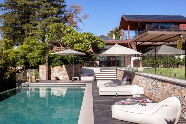 A landscaped outdoor pool terrace beside a multi-level modern home. The rectangular swimming pool is surrounded by dark decking, pale upholstered loungers, umbrellas, stone retaining walls, glass balustrades, and planted greenery. The house features extensive glazing, timber-toned cladding and framing, broad roof overhangs, and a contemporary pitched roof.
