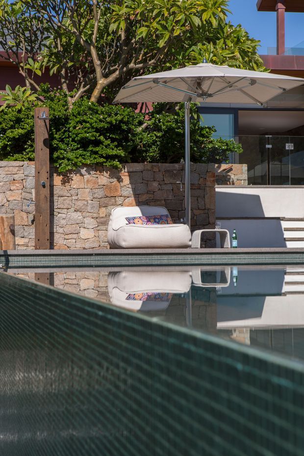 A sunny residential pool area with dark tiled pool walls and reflective water in the foreground. A stone retaining wall, lush tropical planting, outdoor shower, large shade umbrella, lounge chair, and contemporary glazing and steps define the setting. The scene has a relaxed resort-like atmosphere with warm natural stone and greenery alongside modern architectural elements.