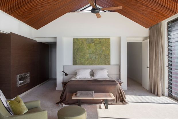 A refined bedroom with a central upholstered bed, dark brown bedding, and a large green-toned artwork above the headboard. The room features a dramatic pitched timber-slat ceiling with an integrated ceiling fan, white walls, dark timber wall panelling with a fireplace, soft grey carpet, full-height curtains, and louvred glazing on the right. Green upholstered lounge chairs and a round ottoman sit in the foreground. Natural light creates a warm, calm atmosphere.