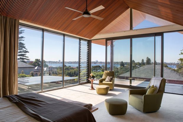 A bright, spacious bedroom with expansive floor-to-ceiling glazing overlooking a leafy waterfront setting. The room features a low-profile bed, sculptural lounge chairs and ottoman, a timber slatted ceiling, a ceiling fan, neutral carpet, full-height curtains, and dark-framed windows with louvred ventilation panels. The interior has a refined mid-century modern character with strong indoor-outdoor connection.