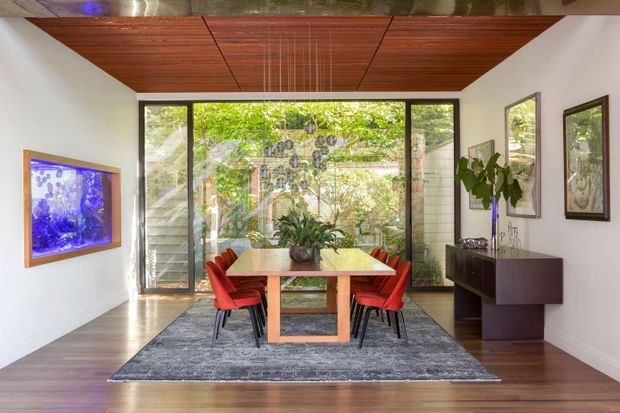 A refined dining room with a timber dining table and bright red upholstered chairs, set on a large grey rug. The room features warm dark timber flooring, a dramatic timber-clad ceiling with recessed linear detailing, white walls, extensive floor-to-ceiling glazing, and a dark built-in sideboard. Framed artwork and a blue-lit wall-mounted aquarium or artwork add visual accents. Lush greenery is visible beyond the glazing.
