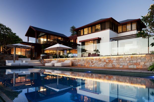 Dusk exterior view of a contemporary multi-level residence arranged around a swimming pool. The home features white rendered walls, dark pitched roof forms, extensive glazing, timber-toned louvres and screens, warm internal lighting, and a substantial natural-stone retaining wall beside the pool. Outdoor seating and umbrellas occupy the terrace, with strong reflections in the water.