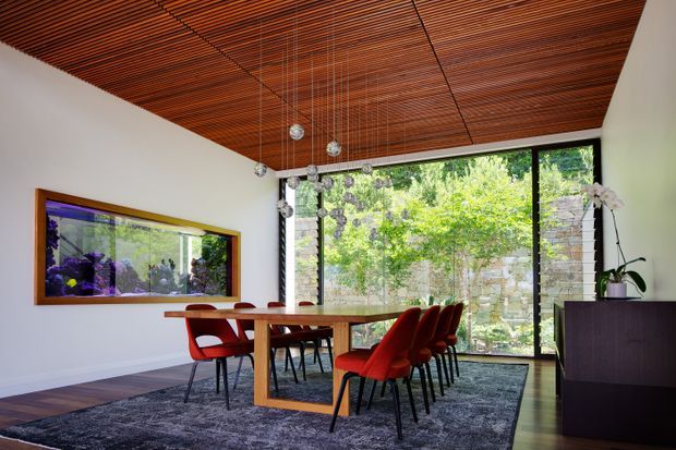 A refined dining room with a long timber dining table surrounded by red upholstered chairs. The room features a dramatic slatted timber ceiling, white walls, dark timber flooring, a large patterned area rug, extensive floor-to-ceiling glazing overlooking a green garden, and a dark built-in sideboard. A long framed aquarium or reflective feature is mounted on the left wall, while pendant lights hang above the table.