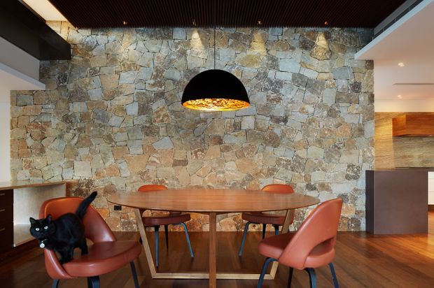 Dining area with a substantial irregular natural-stone feature wall, round timber dining table, sculptural pendant light, and modern leather dining chairs. Dark slatted ceiling, timber flooring, and warm wood cabinetry create a refined contemporary interior. A cat is present in the foreground.