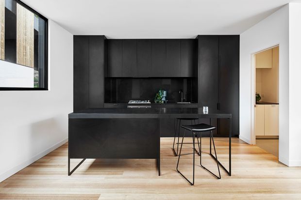 Contemporary minimalist kitchen with a large black built-in cabinetry wall, integrated dark benchtops, and a projecting island with slim metal legs. Pale timber flooring, white walls, a black-framed window, and a small black bar stool create a high-contrast, refined interior. A doorway reveals additional light-wood cabinetry in an adjacent space.