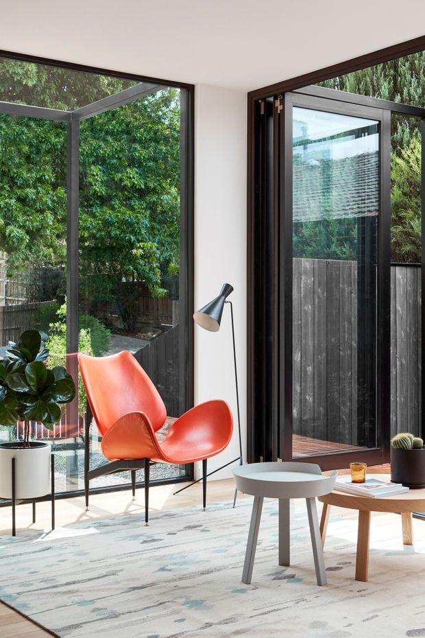 Bright contemporary sitting area with floor-to-ceiling corner glazing and dark-framed hinged doors opening to a timber deck and fenced garden. The room has white walls, pale flooring, a patterned neutral rug, two vivid orange-red sculptural lounge chairs, a slim black floor lamp, a round grey coffee table, a timber side table, and indoor plants. Abundant greenery is visible through the glazing.