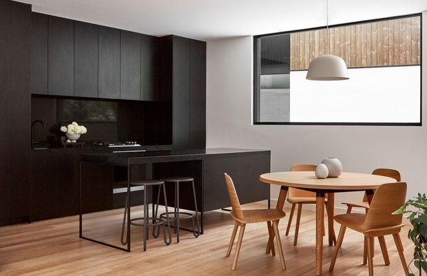 Contemporary open-plan kitchen and dining area with dark, full-height timber cabinetry, a black island and integrated cooktop, light timber flooring, and a round timber dining table with matching mid-century-inspired chairs. A large framed window provides natural light and reveals a timber-clad exterior surface beyond.