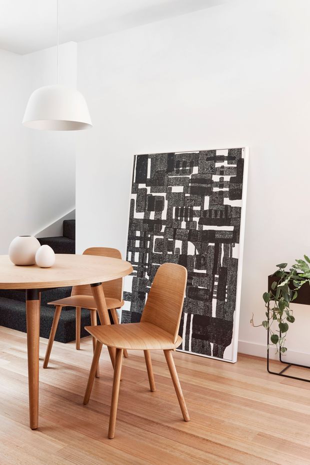 A bright, minimalist dining area with a round timber dining table and matching mid-century-style wooden chairs. The room has white walls and ceiling, pale timber flooring, a sculptural white pendant light, a dark upholstered sofa in the background, and a large black-and-white abstract artwork leaning against the wall. A potted trailing plant adds greenery at the right.