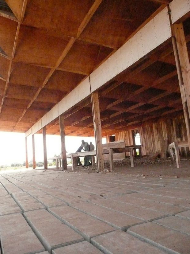 A low-angle view beneath a large rustic timber structure, showing exposed wooden roof framing, posts, beams, weathered timber walls, and a broad paved brick floor. Several people and simple built elements are visible in the background, with bright daylight entering from the open side.