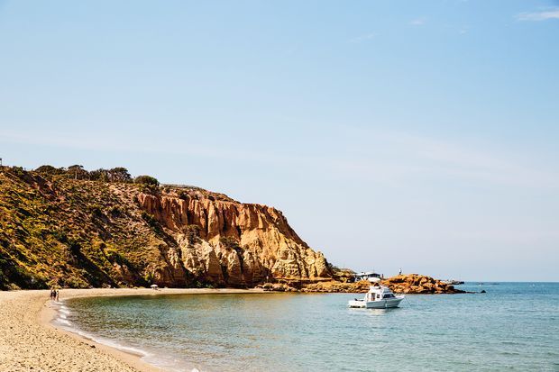 A quiet sandy beach curves around a sheltered turquoise cove, backed by a rugged ochre and cream coastal cliff with low scrub vegetation. A small white motorboat floats near the rocky shoreline under a clear blue sky.