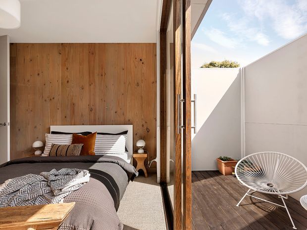 A bright, contemporary bedroom opening directly onto a private timber-decked courtyard or terrace. The room features a full-height natural timber feature wall behind the bed, neutral bedding with patterned and rust-coloured cushions, pale carpet, and simple bedside lighting. Large timber-framed glazed doors connect the bedroom to the sunlit outdoor deck, which has a white rendered privacy wall, potted plant, and wire lounge chair.