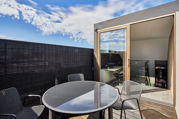 A contemporary outdoor terrace or balcony with a round glass-top dining table and metal chairs, enclosed by a dark privacy screen. Large sliding glass doors open into a compact home office or study, with a desk, office chair, shelving, and tiled flooring visible inside. The building has a simple modern form with neutral rendered walls and timber-toned door framing, set beneath a bright blue sky.