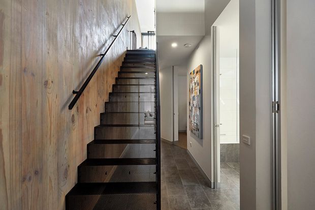 Interior view of a contemporary hallway and stairway. A straight flight of open-riser stairs with dark perforated metal treads and a simple black handrail runs alongside a full-height natural timber wall. The corridor has grey tiled flooring, white walls, recessed ceiling lighting, and open doorways leading to adjacent rooms, including a partially visible tiled bathroom.