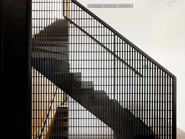 Interior view of a modern stairway enclosed by a dramatic black metal grille balustrade. The straight-run staircase has dark treads and a strong geometric diagonal handrail, with white walls and a discreet linear air vent behind it. Warm timber-toned elements are partially visible beneath the stairs.