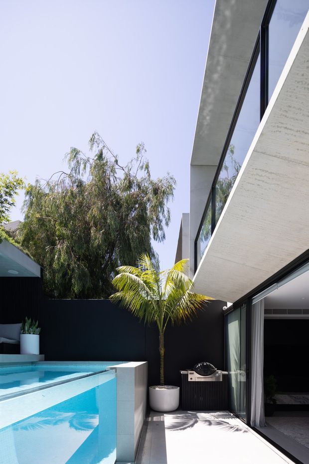 A bright contemporary residential courtyard viewed from an outdoor terrace, featuring a turquoise swimming pool, dark boundary walls, tropical planting, and a sharply cantilevered concrete roof or canopy with large black-framed glazed openings. A compact barbecue sits beside the glazing, with an interior room visible beyond.