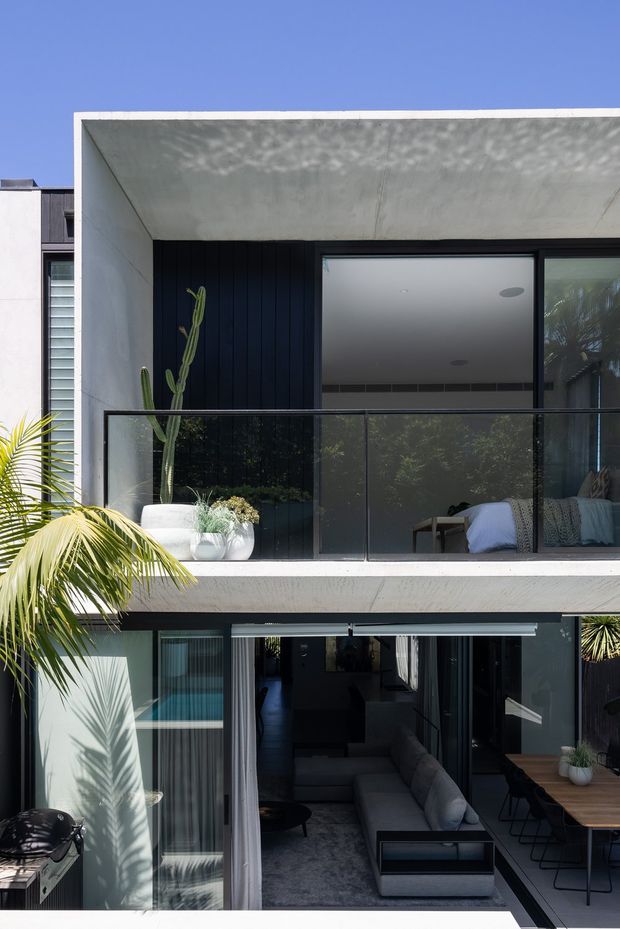 A contemporary multi-storey residential facade with a deep concrete-framed balcony and broad sliding glazing. The upper balcony has a frameless-style dark glass balustrade, black vertical wall cladding, potted cacti, and a partially visible bedroom. Below, large glazed openings connect a dark, modern living room to an outdoor dining deck, with tropical planting and strong sunlight creating a relaxed indoor-outdoor setting.