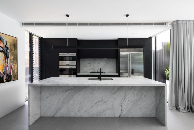 Contemporary open-plan kitchen viewed straight on, centred on a large waterfall-edge marble island with an integrated sink and black tapware. Dark matte-black full-height cabinetry forms the rear wall, incorporating stacked appliances, open shelving, a marble splashback and concealed cooking area. Floor-to-ceiling glazing and sheer grey curtains appear at the right, with light walls, polished concrete-look flooring and minimal recessed lighting creating a refined monochromatic interior.