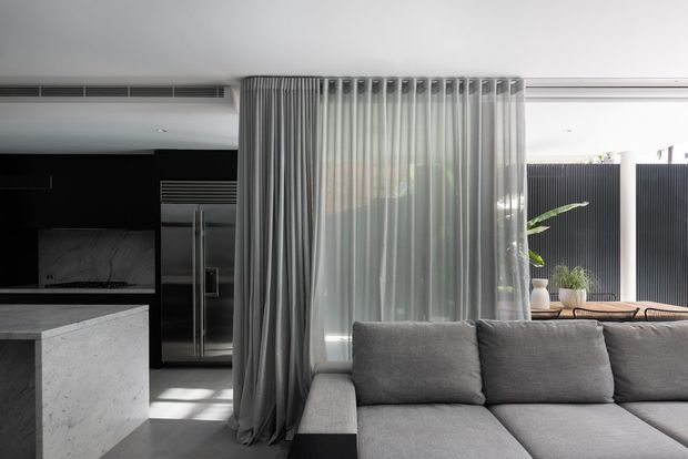 A refined open-plan interior combining a dark kitchen with stainless-steel appliances and a pale stone island, alongside a large grey sectional sofa. Floor-to-ceiling sheer curtains create a soft divider between the living and kitchen areas, with a partially visible planted courtyard beyond. The space has a restrained monochromatic palette, clean lines, and abundant filtered daylight.