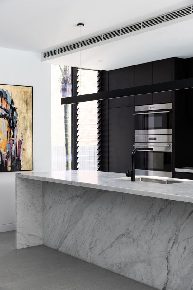 A bright contemporary kitchen dominated by a large waterfall island clad in veined pale stone. The island incorporates a black gooseneck tap and undermount sink, while the rear wall features full-height matte-black cabinetry with integrated stainless-steel ovens and microwave. A long black linear pendant hangs above the island, with a high-level linear air-conditioning grille and louvred window providing architectural detail. Pale grey flooring and white walls balance the dark joinery, while an abstract artwork adds colour at the side.