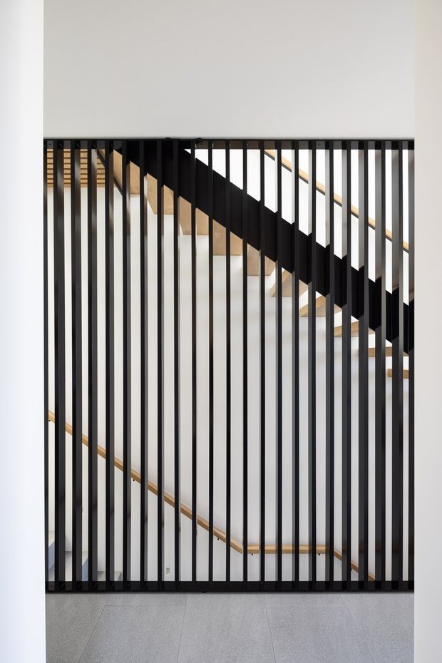 A minimalist interior view of a stairwell seen through a full-height black vertical metal balustrade or screen. The staircase has light timber treads and handrails, white walls, and a grey tiled floor. Strong geometric lines and contrasting black, white, and timber elements create a clean contemporary architectural composition.