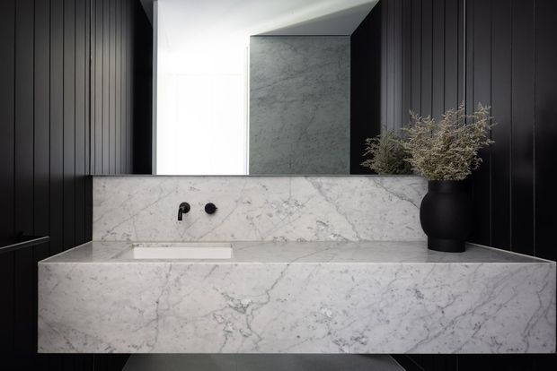 A sophisticated contemporary bathroom vanity featuring a floating rectangular white marble basin and backsplash with prominent grey veining. Matte black vertically fluted wall panelling surrounds the vanity, while a large mirror reflects a dark stone-clad wall and bright ceiling. Minimal black wall-mounted tapware and a matte black vessel with dried branches provide restrained contrast.