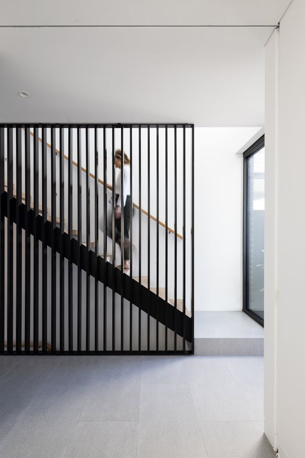 A bright contemporary interior view focused on a staircase enclosed by a striking black vertical-slat balustrade and screen. Pale large-format floor tiles, white walls and ceiling, recessed lighting, and a tall dark-framed window create a clean minimalist architectural setting. A person is visible ascending the stair behind the screen.