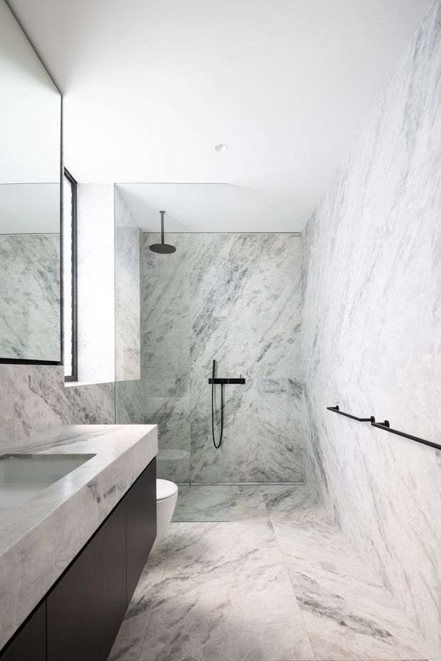 Bright contemporary bathroom with a walk-in glass shower, large-format grey-veined stone wall and floor surfaces, a white stone vanity benchtop with dark fixed cabinetry, frameless mirror, and matte black shower and grab hardware. The space is narrow, clean-lined, and highly monochromatic.