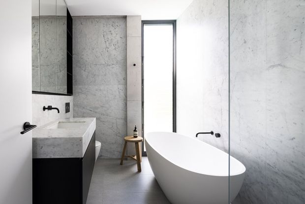A bright contemporary bathroom with a freestanding white bathtub beside a full-height glass shower screen, a concrete-look stone vanity with dark cabinetry, large pale grey marble-effect wall surfaces, and matte black tapware. A tall narrow window provides natural light.