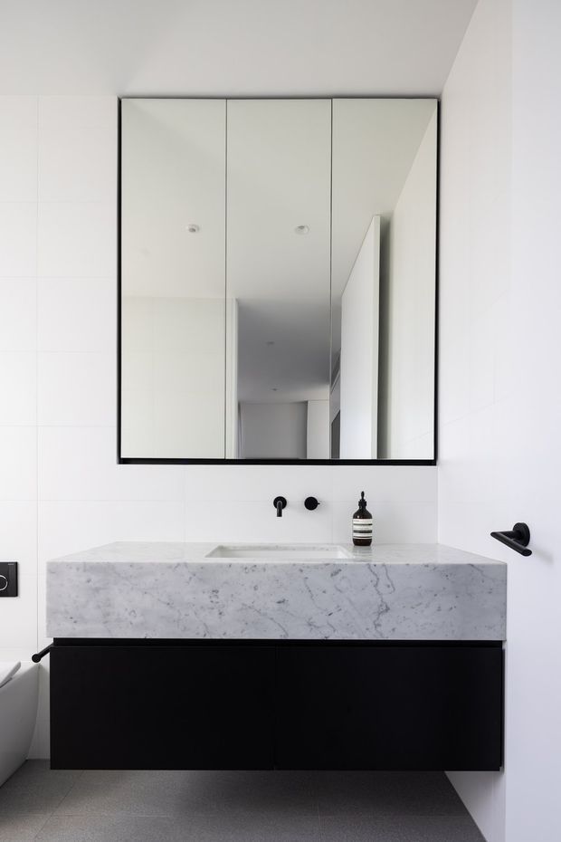 A minimalist contemporary bathroom vanity with a thick white-grey veined stone benchtop and integrated basin, matte black floating cabinetry, black wall-mounted tapware and hardware, and a large black-framed mirrored cabinet. White tiled walls and grey floor tiles create a clean monochromatic scheme.