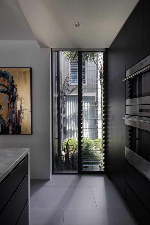 A narrow, contemporary galley kitchen with dark full-height cabinetry and integrated double ovens along the right wall. A dark island with a light grey stone benchtop is partially visible on the left. Large floor-to-ceiling glazing with horizontal louvres opens the kitchen toward a planted courtyard, bringing in strong natural light. The space has pale large-format floor tiles, white walls and ceiling, a dark window frame, and a large abstract artwork.