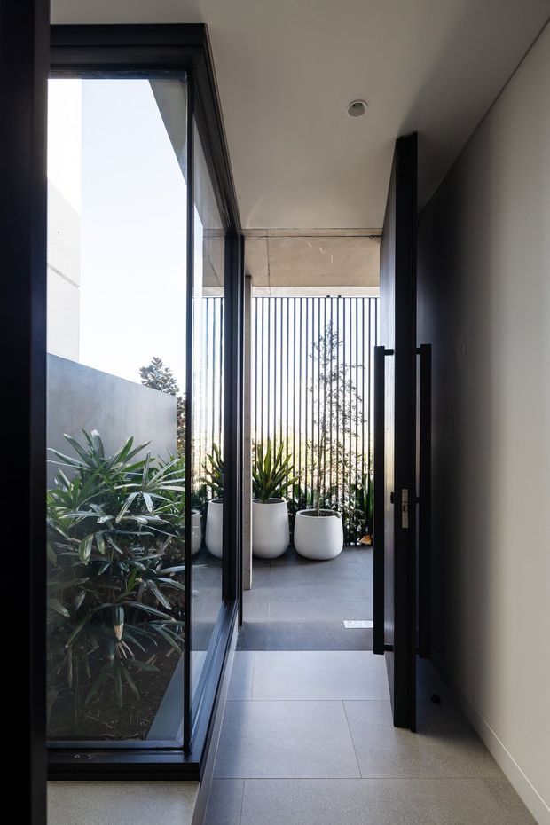 A narrow contemporary entryway opens onto a covered outdoor terrace or courtyard. The space features large black-framed glazed doors and windows, pale smooth walls, a recessed ceiling light, and large-format grey floor tiles. White cylindrical planters with leafy greenery sit beyond the opening, backed by a vertical metal privacy screen. The view is bright and minimalist with strong indoor–outdoor connection.