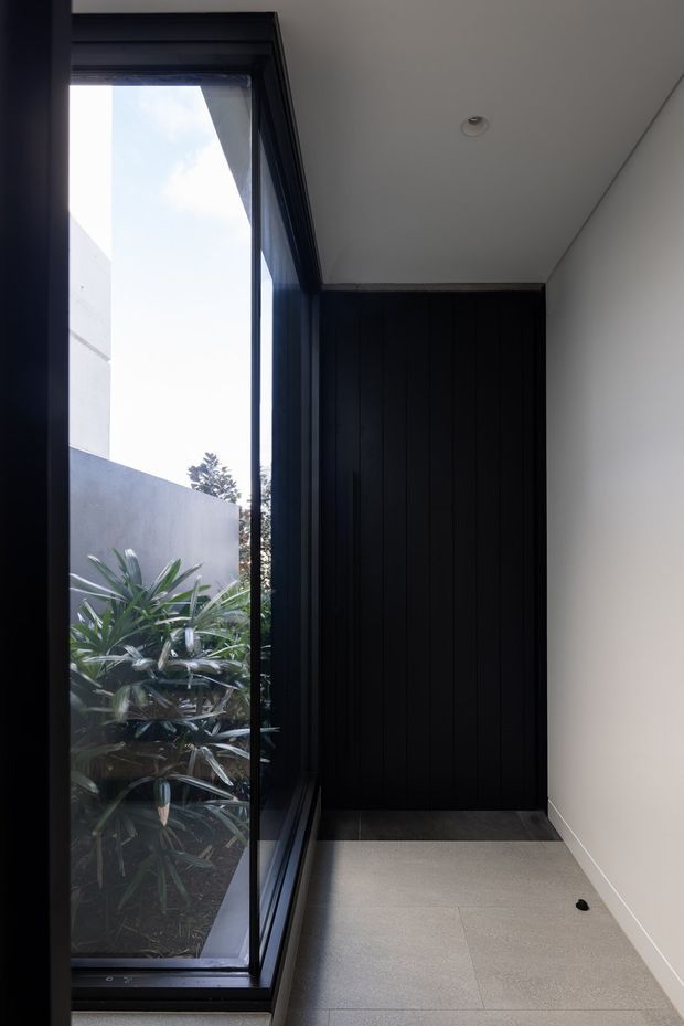 A narrow, minimalist interior passage or entry area with a full-height black-framed glazed wall on the left, overlooking planted landscaping. The rear wall features tall dark vertical-panel joinery or cladding, while the opposite wall and ceiling are plain white. Large pale grey floor tiles and recessed ceiling lighting create a clean, contemporary feel.